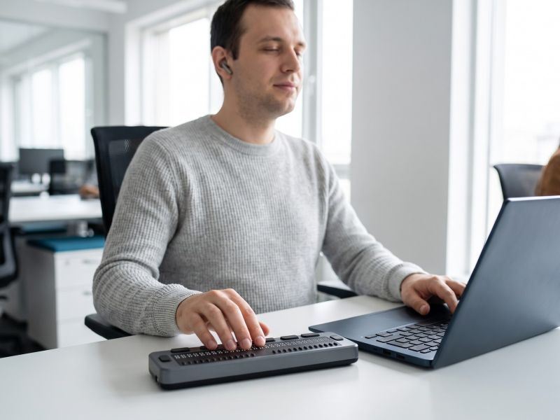 Blind man reading a refreshable braille display beside his laptop while using a screen reader